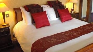 Una cama bien hecha con una colcha roja y varias almohadas, incluidas rojas y marrones. Hay dos lámparas de noche a ambos lados de la cama.