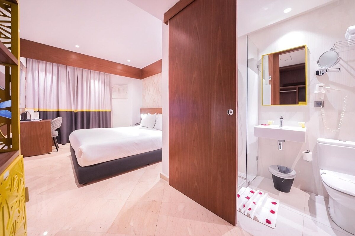 A hotel room with a bed, a desk, a bathroom with a mirror, and a shower.