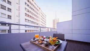 Een balkon met een houten tafel, gedekt voor het ontbijt, inclusief een fruitsalade, gebak en een drankje.