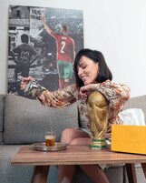 Una persona sirve una bebida en un vaso sobre una mesa de madera, con una estatua dorada y un cartel deportivo enmarcado al fondo.