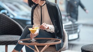 Iemand zit aan een ronde tafel met een drankje, gekleed in een zwart jack over een beige top, in een openluchtomgeving met wazige auto's en een passerende persoon op de achtergrond.