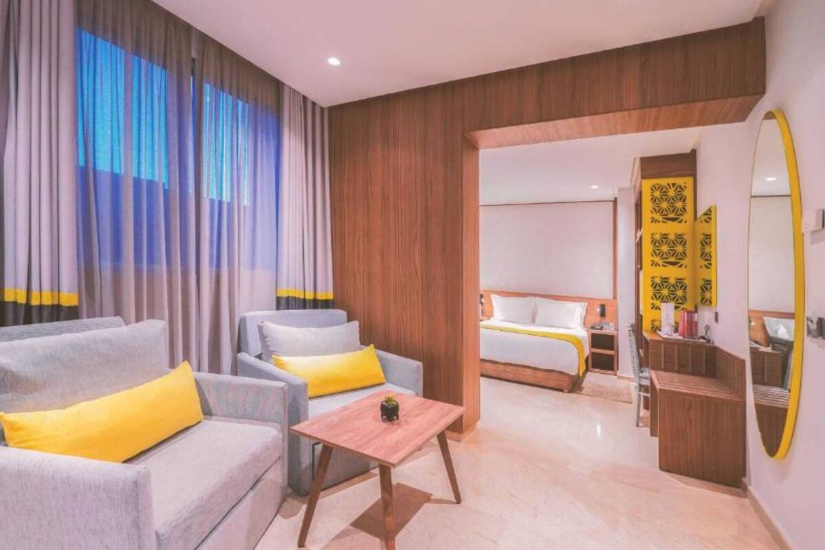 Een hotelkamer met een bank, fauteuil en een klein tafeltje. Er staat een bed met een wit hoofdeinde en een gele accentkleur. De kamer heeft houten lambrisering en een grote spiegel aan de muur.