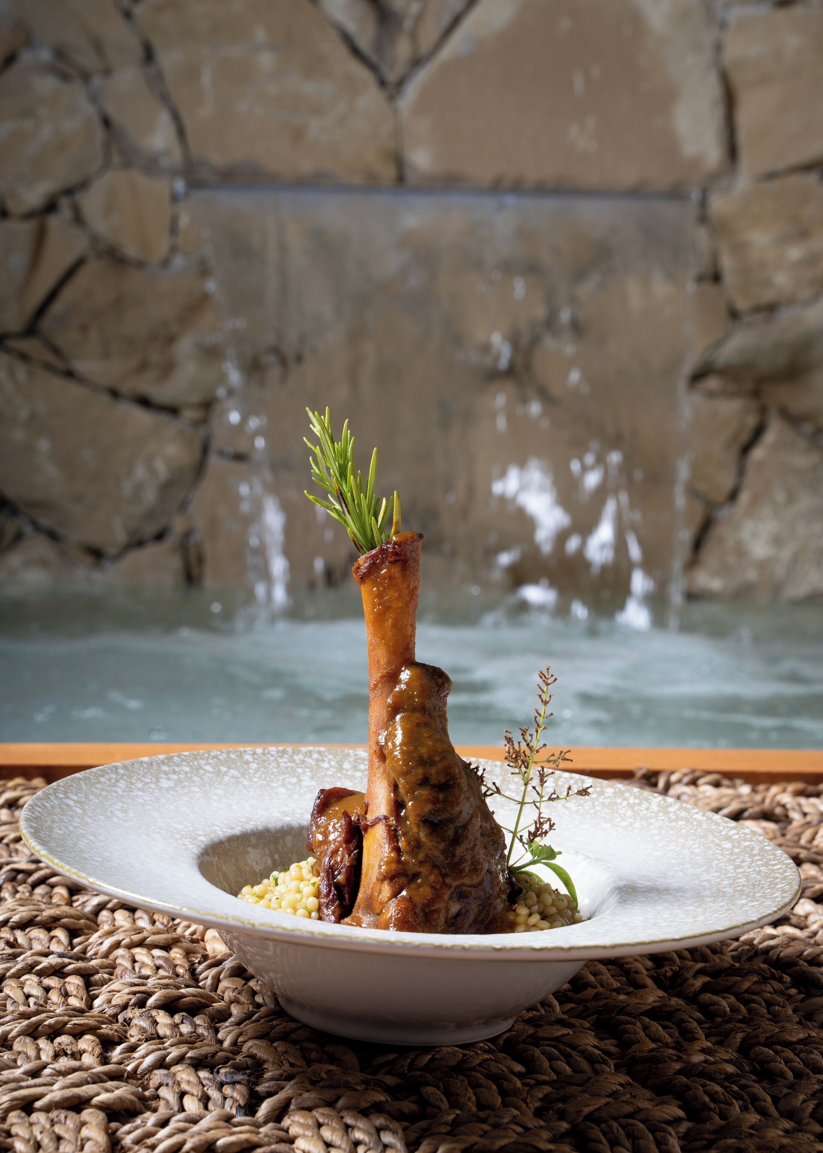 Un piatto con un osso e verdure su una tovaglia lavorata, con un muro in pietra e una fontana sullo sfondo.