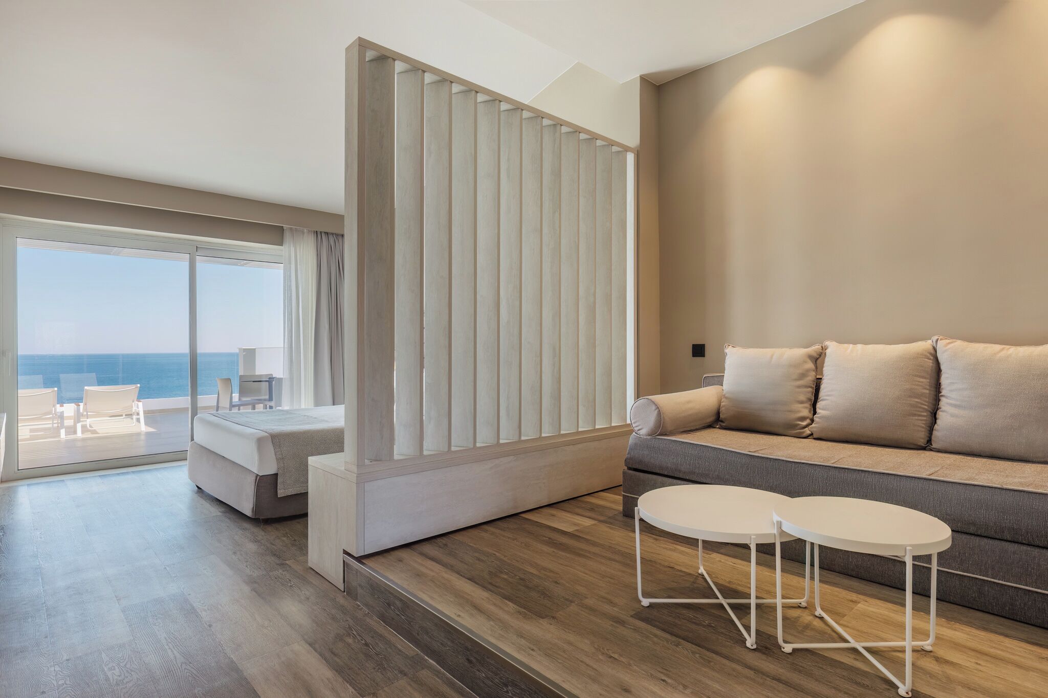 A modern hotel room with a large sliding glass door leading to a balcony, a sofa with cushions, and two small white tables.