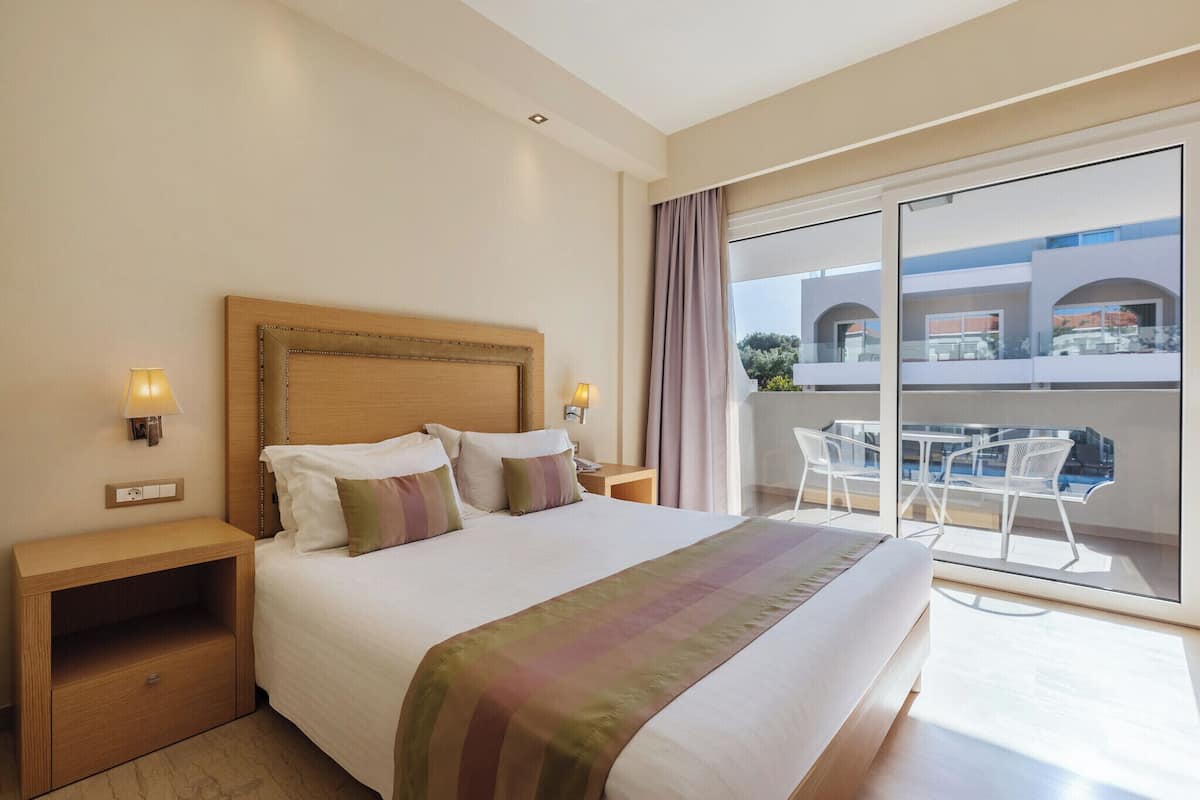A hotel room with a large bed, two bedside tables with lamps, a balcony with white chairs, and a view of the building's exterior.