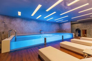 Una piscina interior moderna con un elemento acuático iluminado en azul, suelo de madera y sillones blancos para lounge.