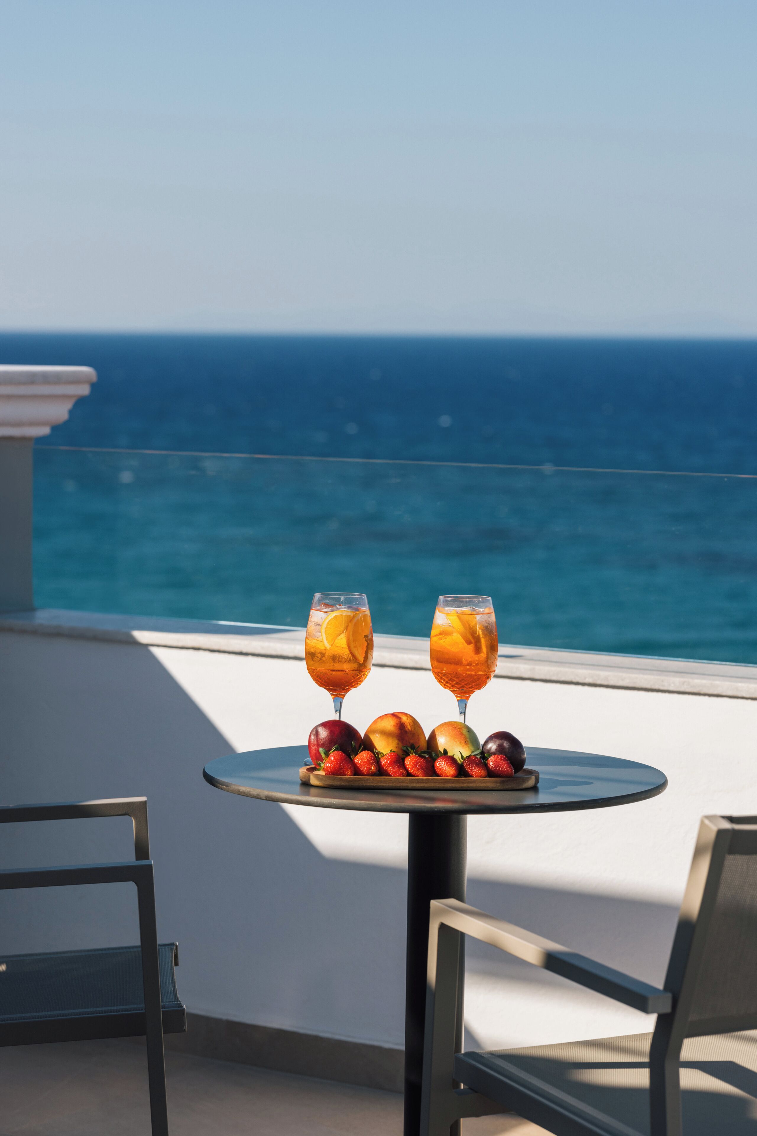 A table with two glasses of orange juice and a plate of fruit on a balcony overlooking the sea.