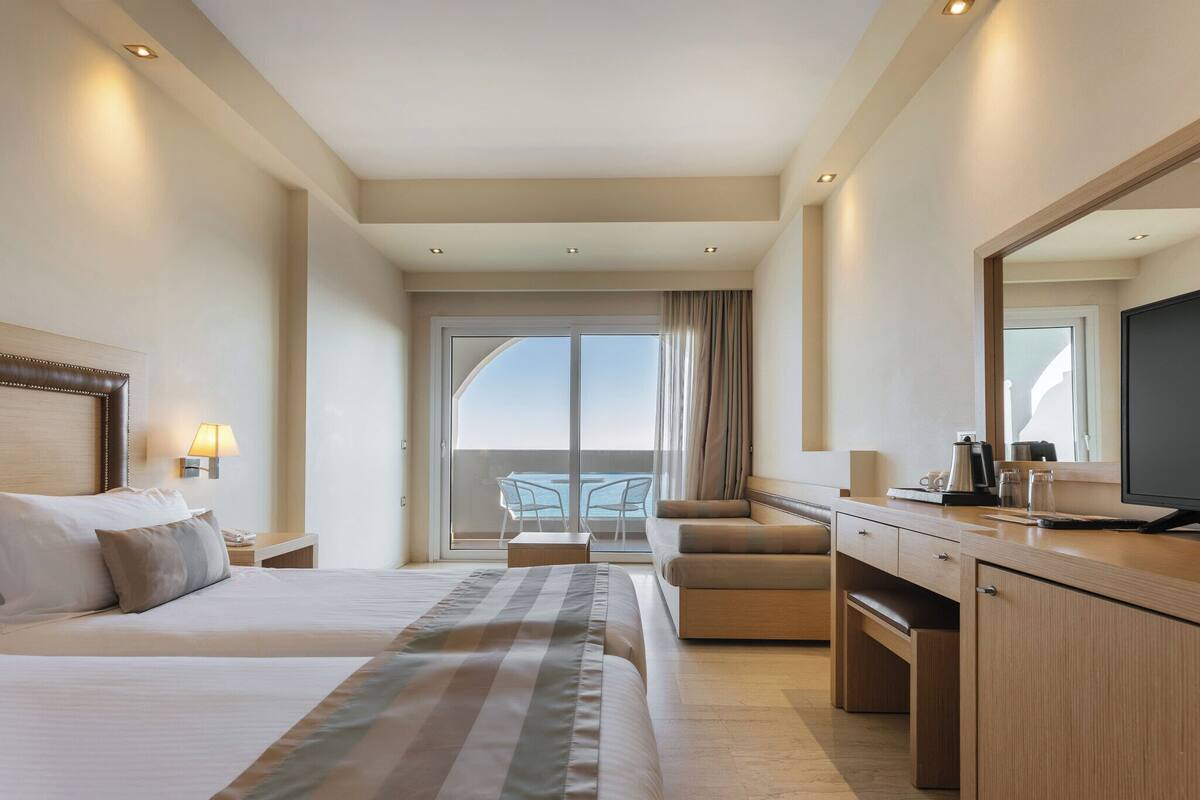A hotel room with a large bed, a desk, a chair, a TV, and a balcony with a view.