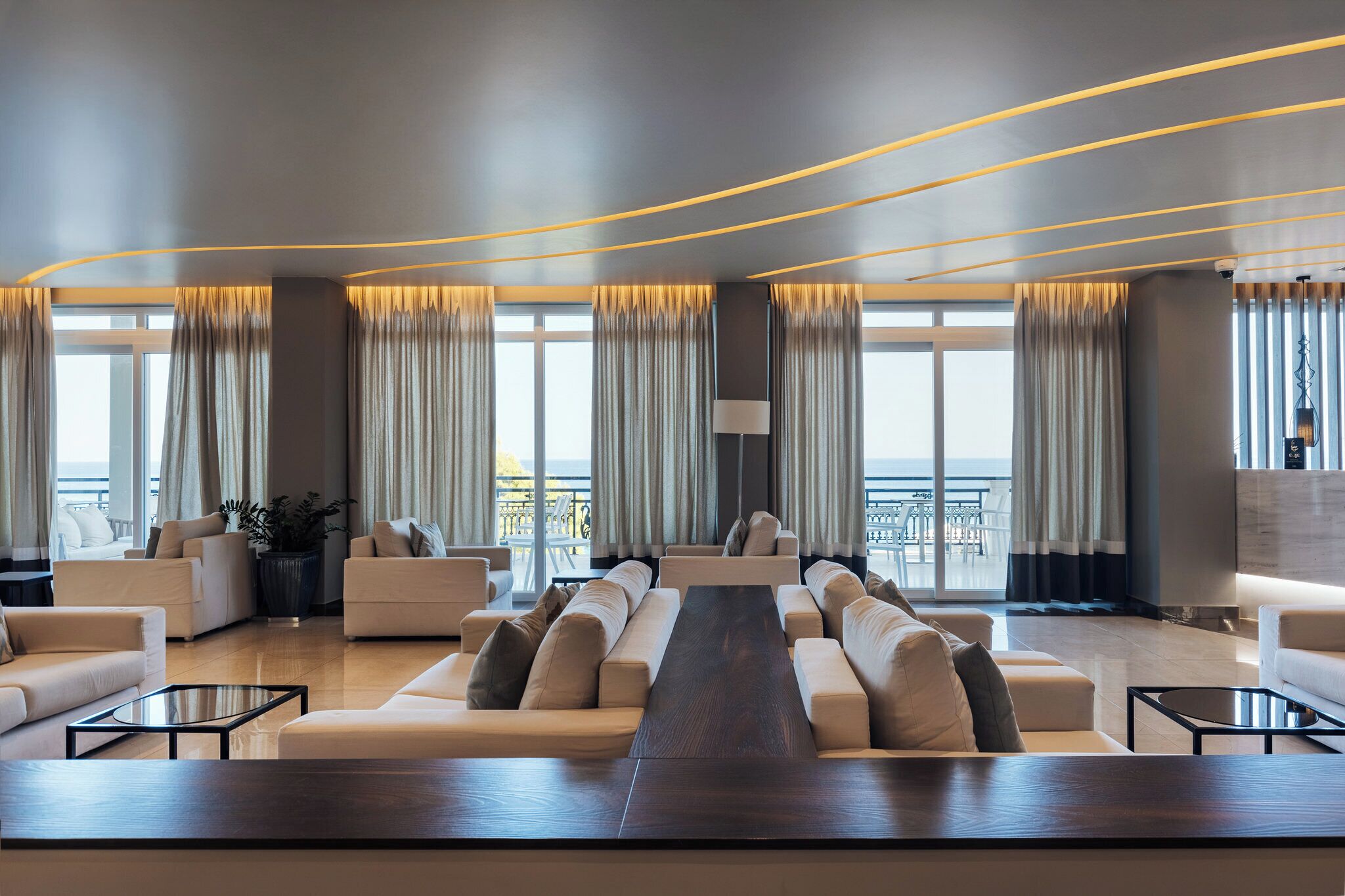 A modern living room with a long wooden table, sofas, and large windows with curtains.