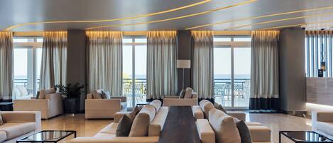 A modern living room with a long wooden table, sofas, and large windows with curtains.
