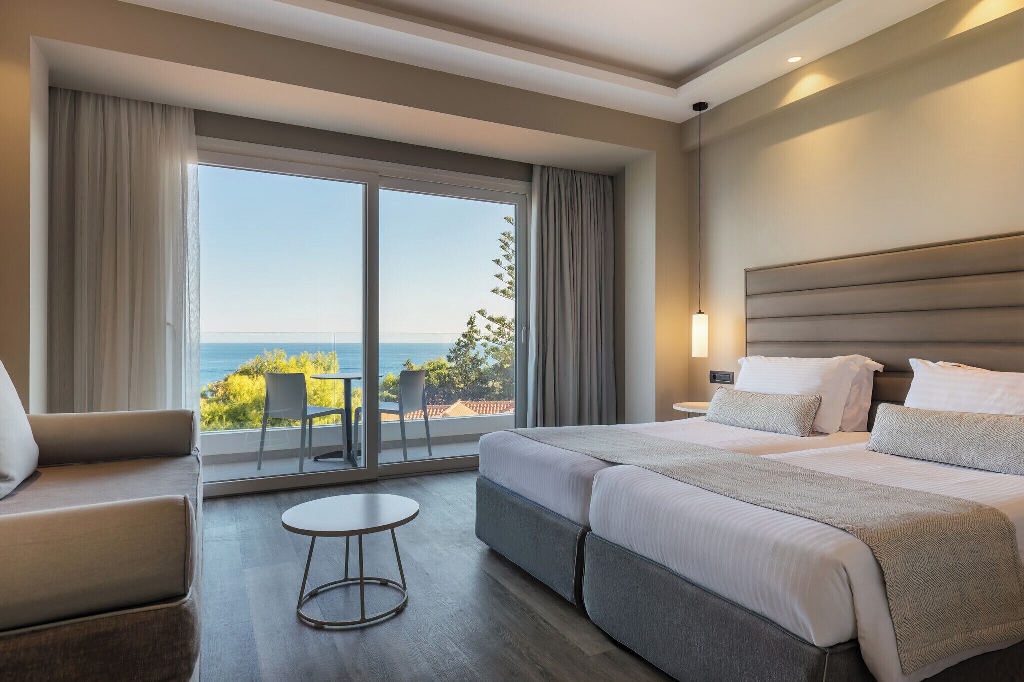 Habitación de hotel con un gran ventanal corredera que da a un balcón con vistas al mar. Se ve una cama con dos almohadas y una mesita de noche.