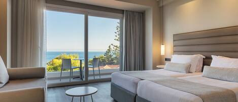 Habitación de hotel con un gran ventanal corredera que da a un balcón con vistas al mar. Se ve una cama con dos almohadas y una mesita de noche.