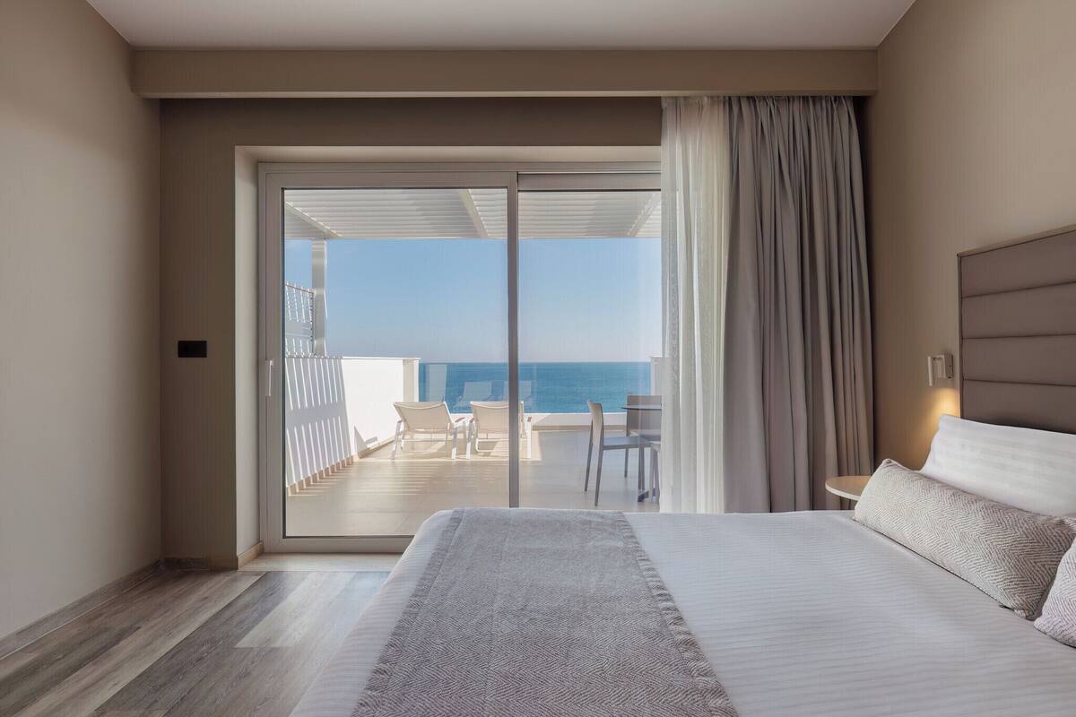 A hotel room with a large bed, a view of the ocean, and a balcony with chairs.