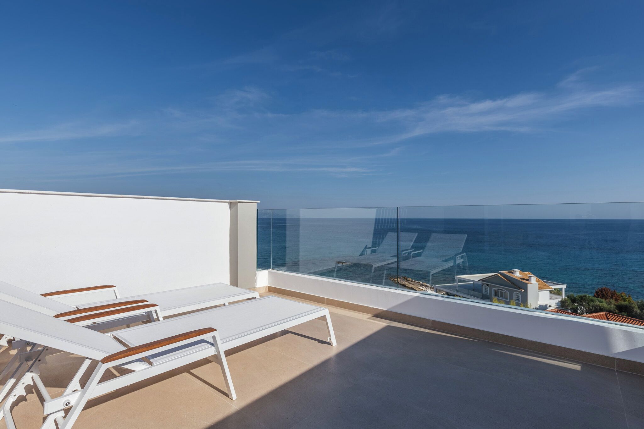 A balcony with white lounge chairs, a glass railing, and a view of the sea and buildings.