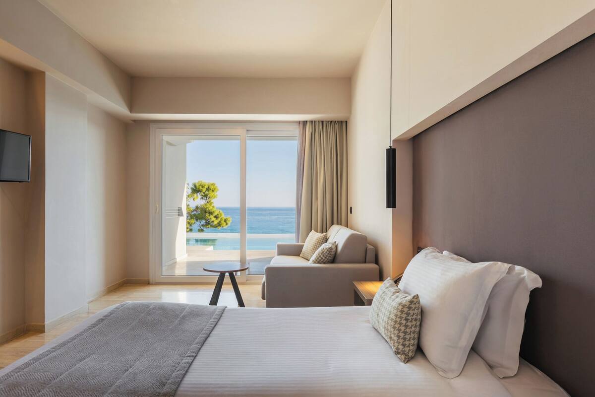A hotel room with a bed, a sofa, a small table, and a TV. There is a balcony with a view of the ocean.