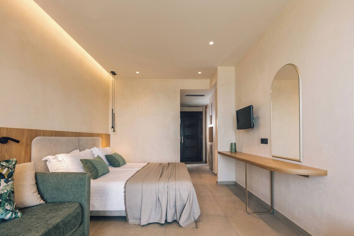 A modern hotel room with a bed, a sofa, a TV, and a mirror.