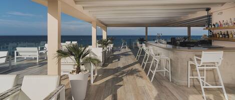 A rooftop bar with white bar stools, a large potted plant, and a view of the ocean.