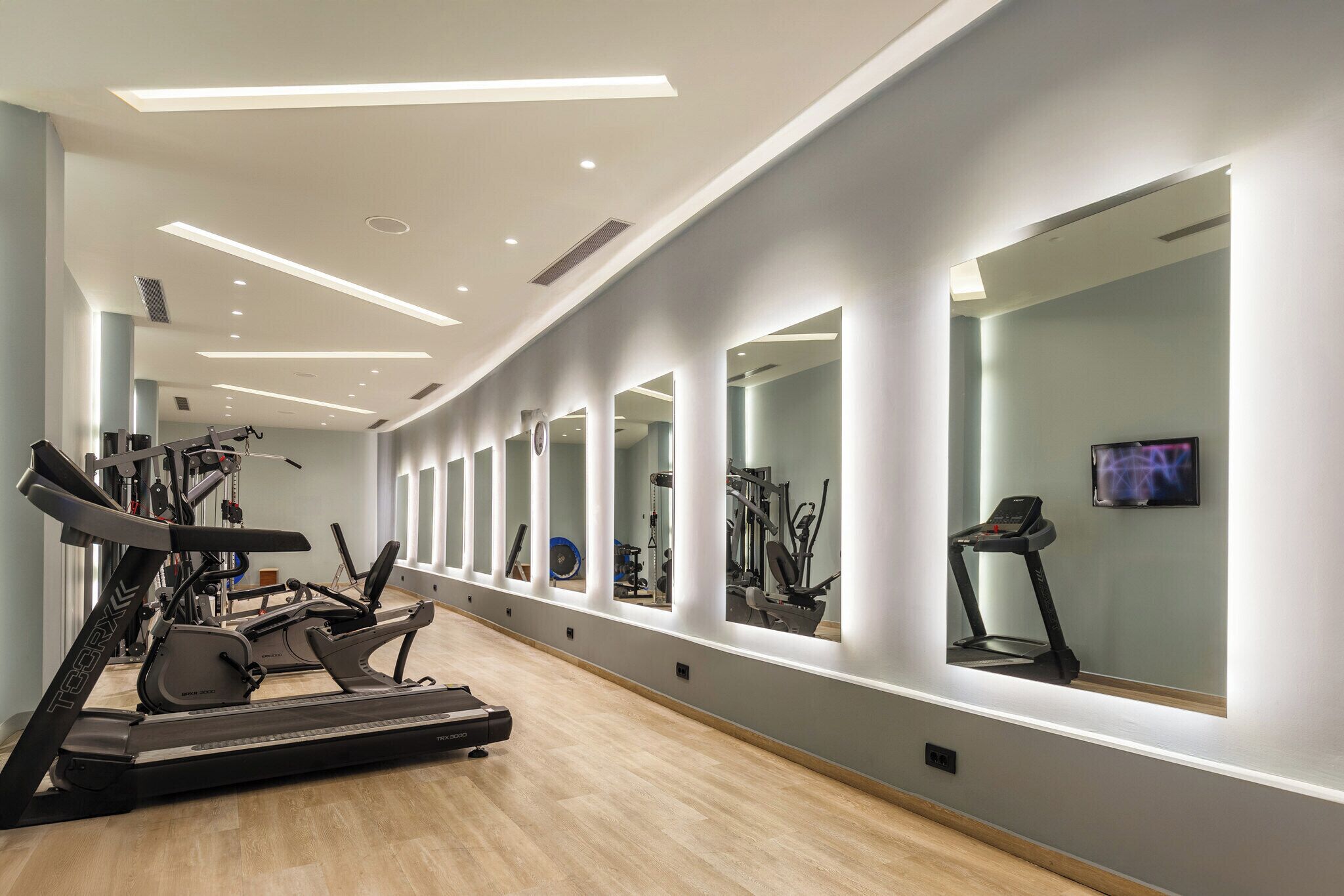 A modern gym with cardio equipment, mirrors, and a TV screen.