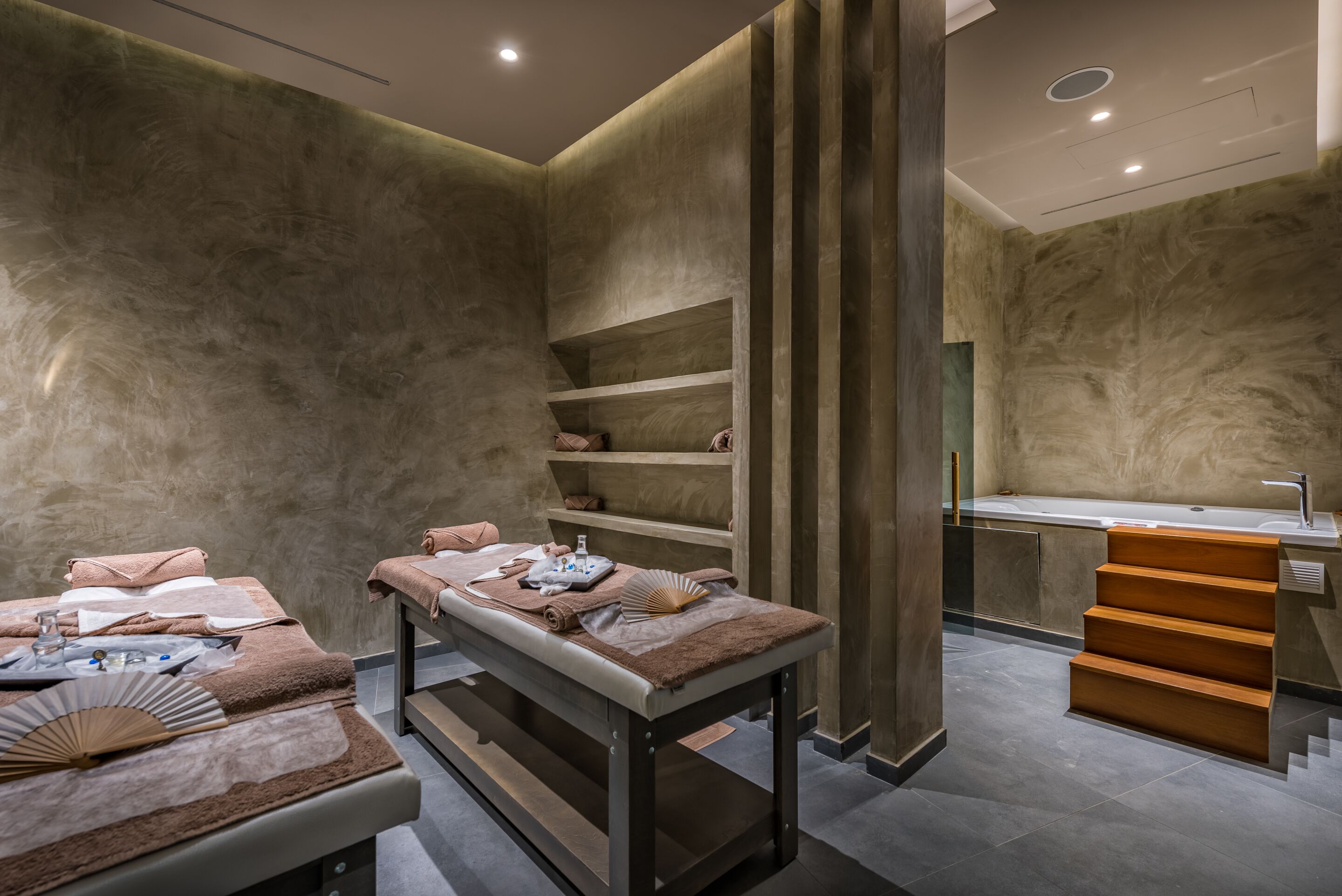 Un spa moderno con una camilla de masaje, estantes con toallas y una escalera de madera que conduce a un baño con lavamanos.