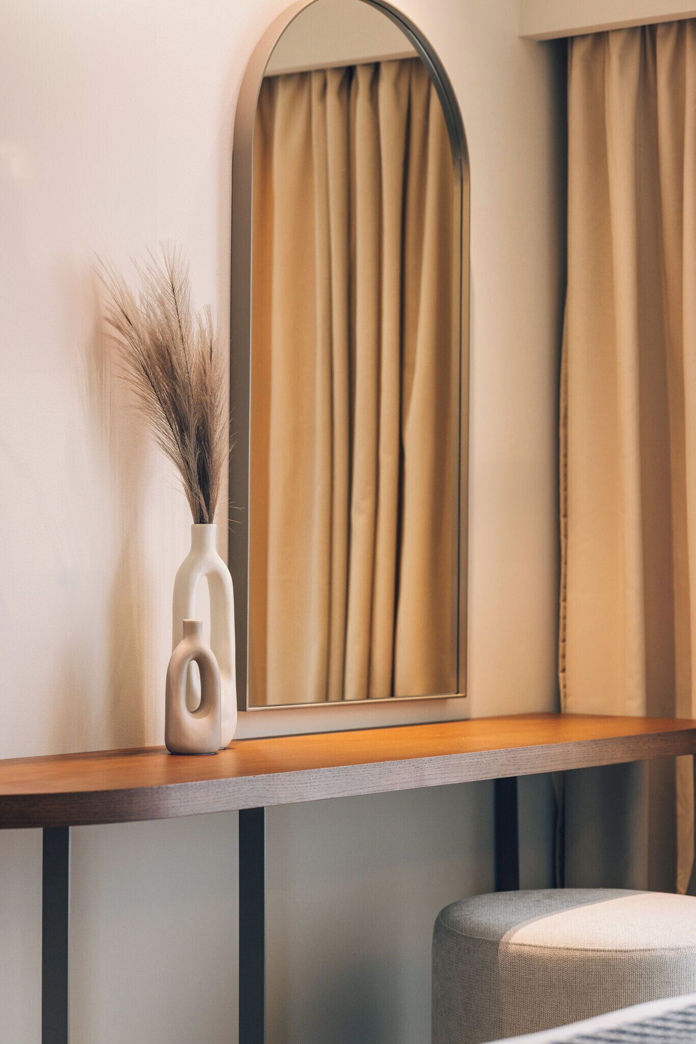 A modern interior with a wooden console table, a vase with dried plants, and a mirror reflecting curtains.