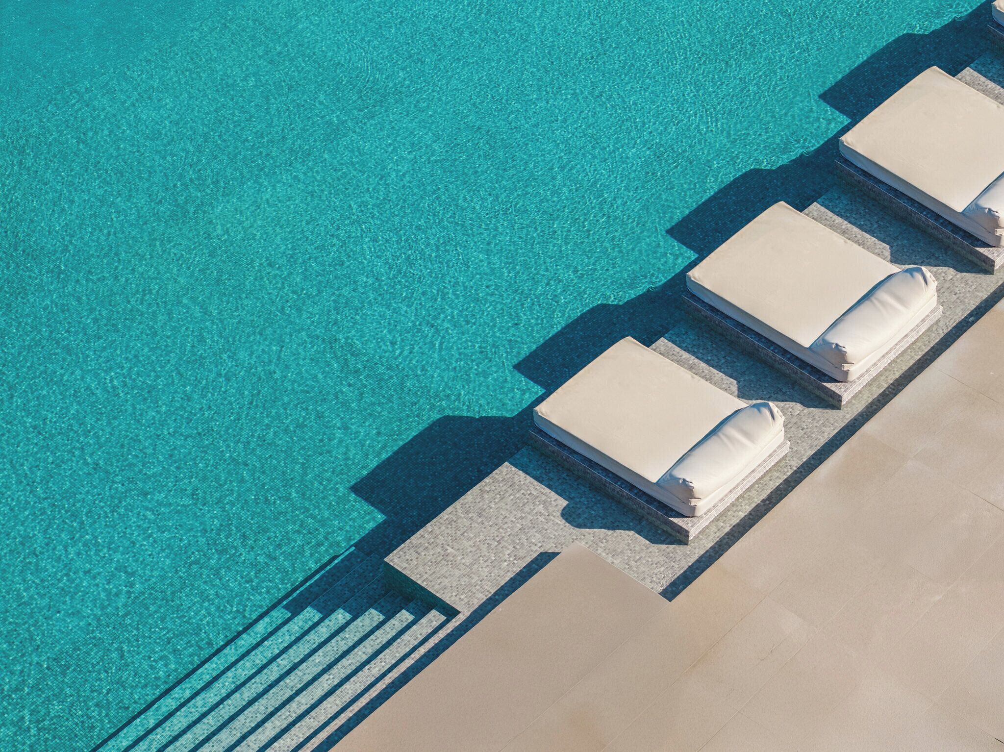 An aerial view of a pool area with clear blue water, tiled steps leading to lounge chairs with white cushions.