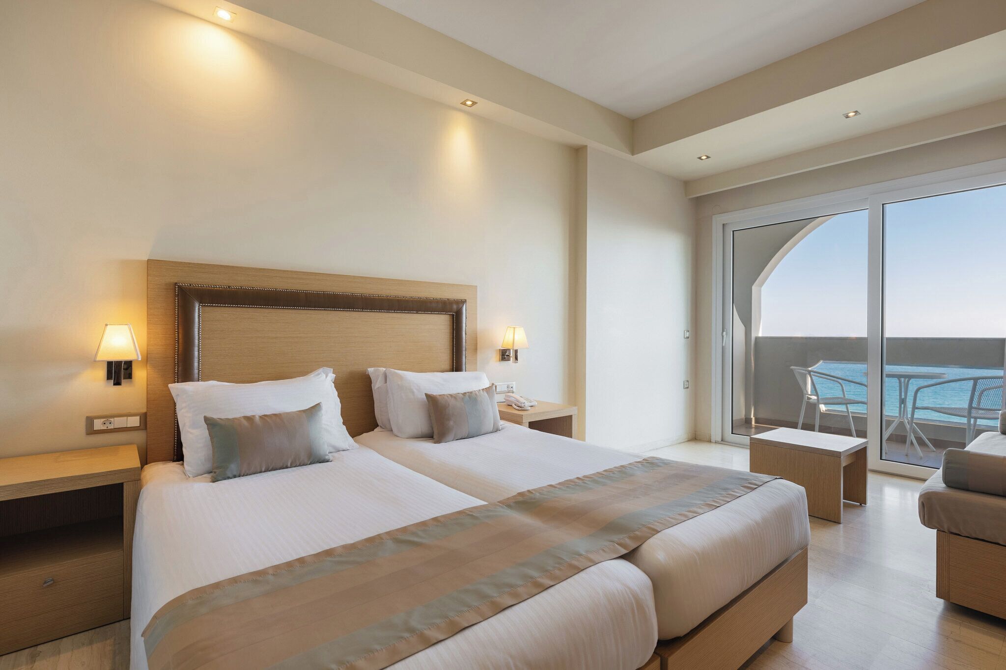 A hotel room with two beds, a balcony with a table and chairs, and a view of the sea.