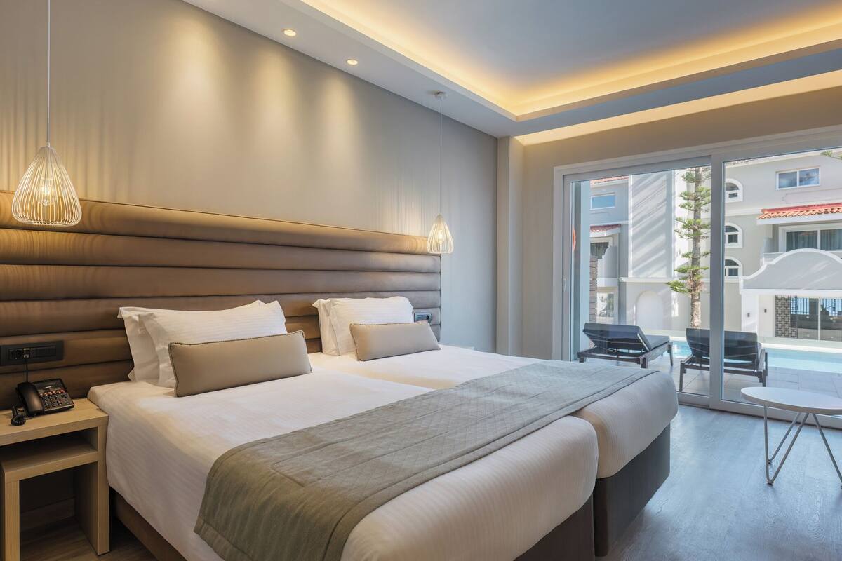 A hotel room with two beds, a bedside table with a phone, a small table with chairs, and a large window with a view of buildings.