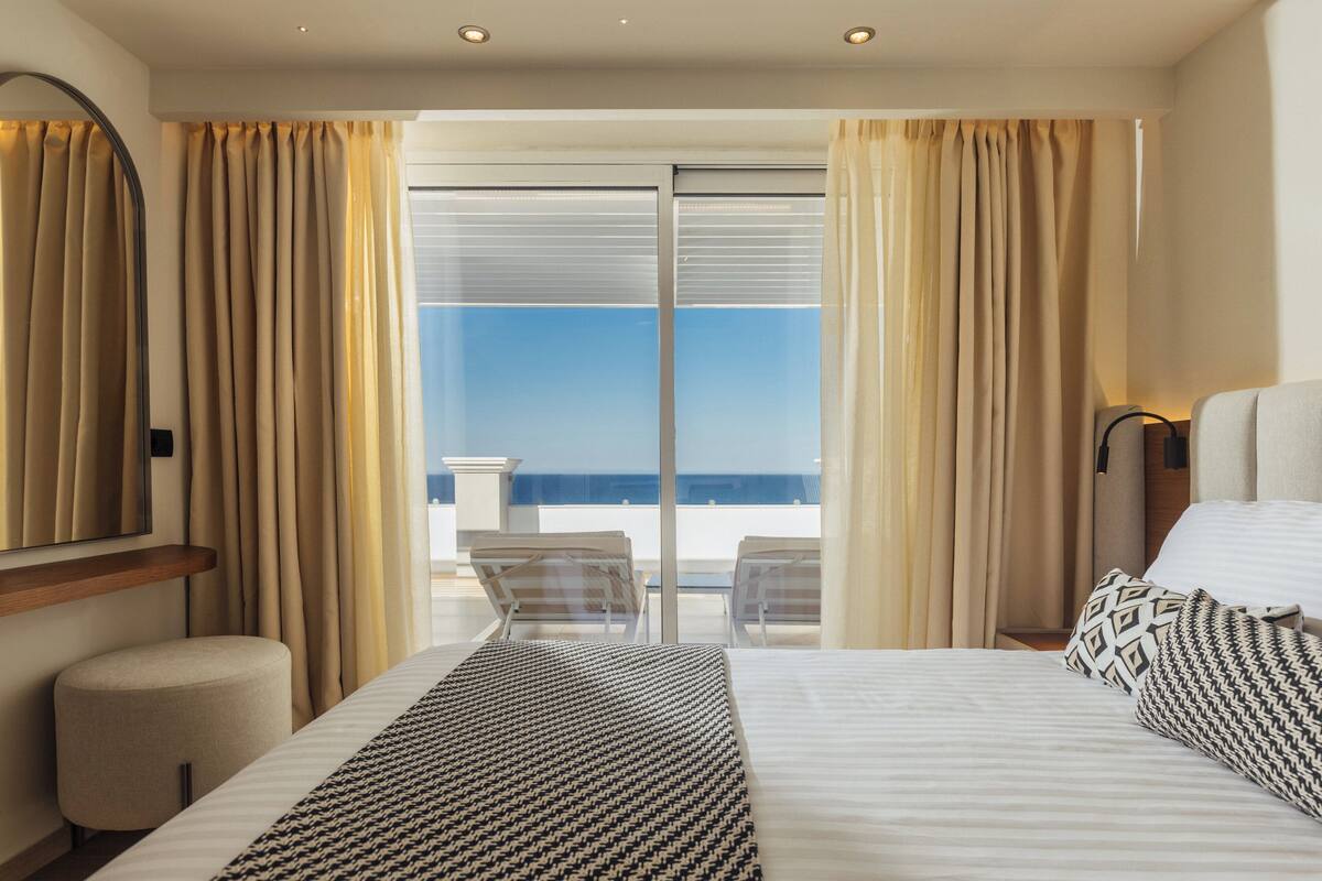 A hotel room with a bed, a chair, a small table, and a view of the sea through large windows.
