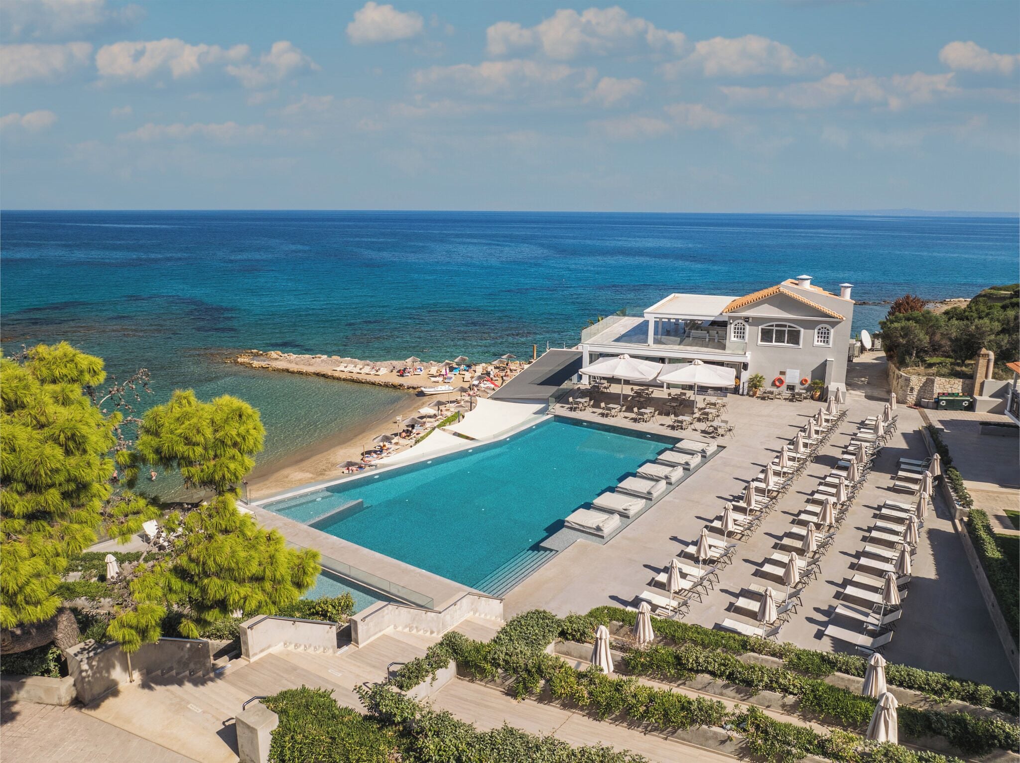 Vista aérea de un complejo turístico con piscina, sillones de descanso y vista al mar.