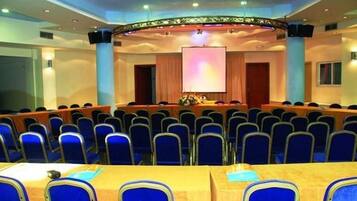 A conference room with rows of blue chairs, a stage with a podium, and a large screen.