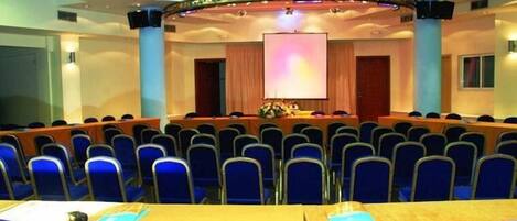 A conference room with rows of blue chairs, a stage with a podium, and a large screen.