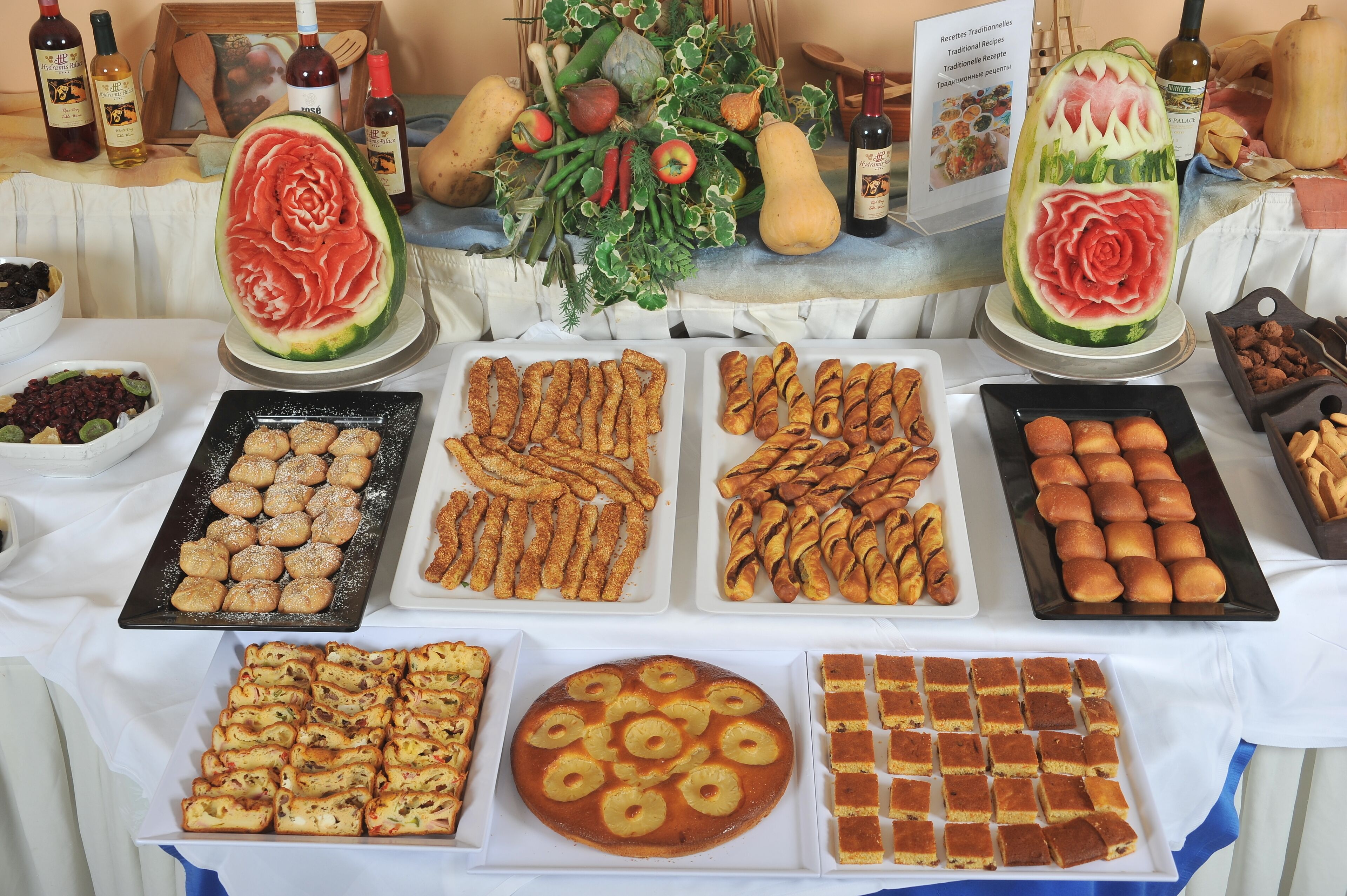 Mesa de buffet com diversos alimentos, incluindo doces, pão e uma melancia com uma rosa esculpida.