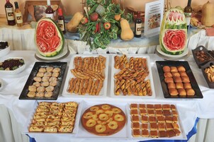 Mesa de buffet com diversos alimentos, incluindo doces, pão e uma melancia com uma rosa esculpida.