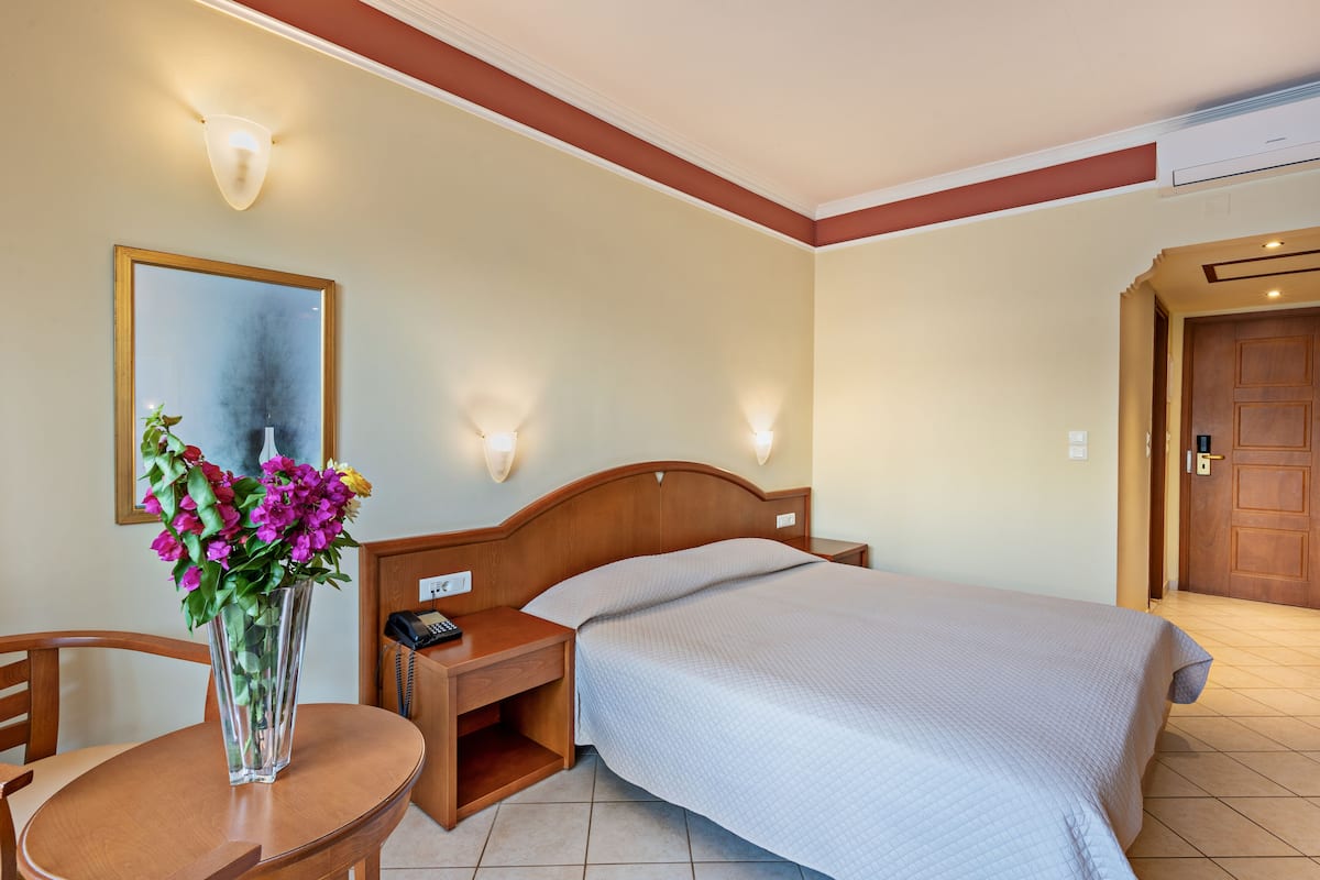 A hotel room with a bed, bedside table, chair, and a vase with flowers.