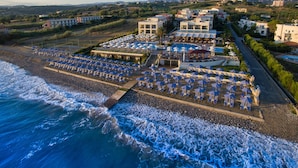 An aerial view of a coastal hotel with multiple beachside umbrellas, a swimming pool, and a mountain backdrop. - Hydramis Palace Beach Resort (Apokoronas)