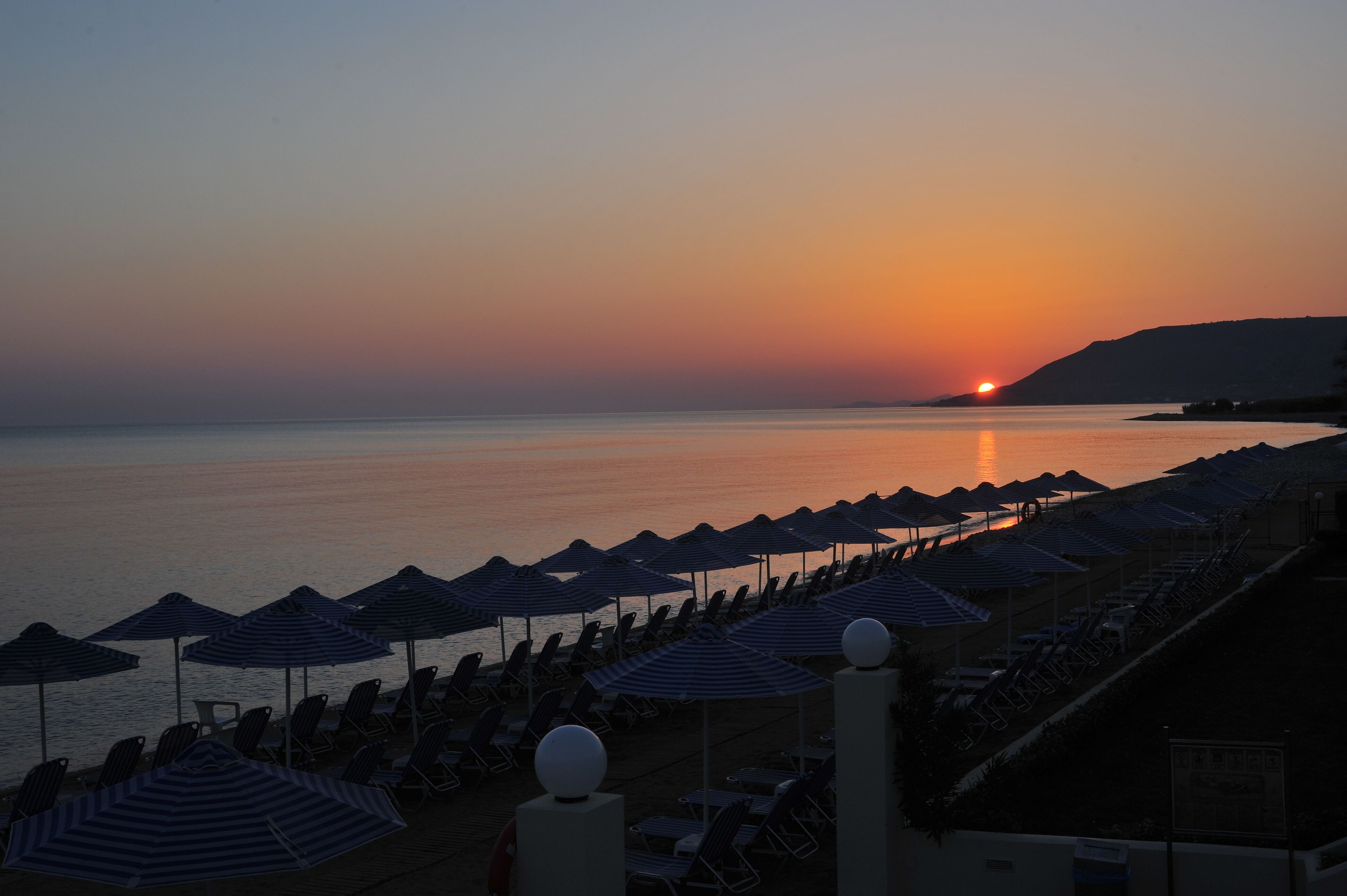 Uma praia com fileiras de espreguiçadeiras e guarda-sóis, um pôr do sol ao fundo e mar calmo.