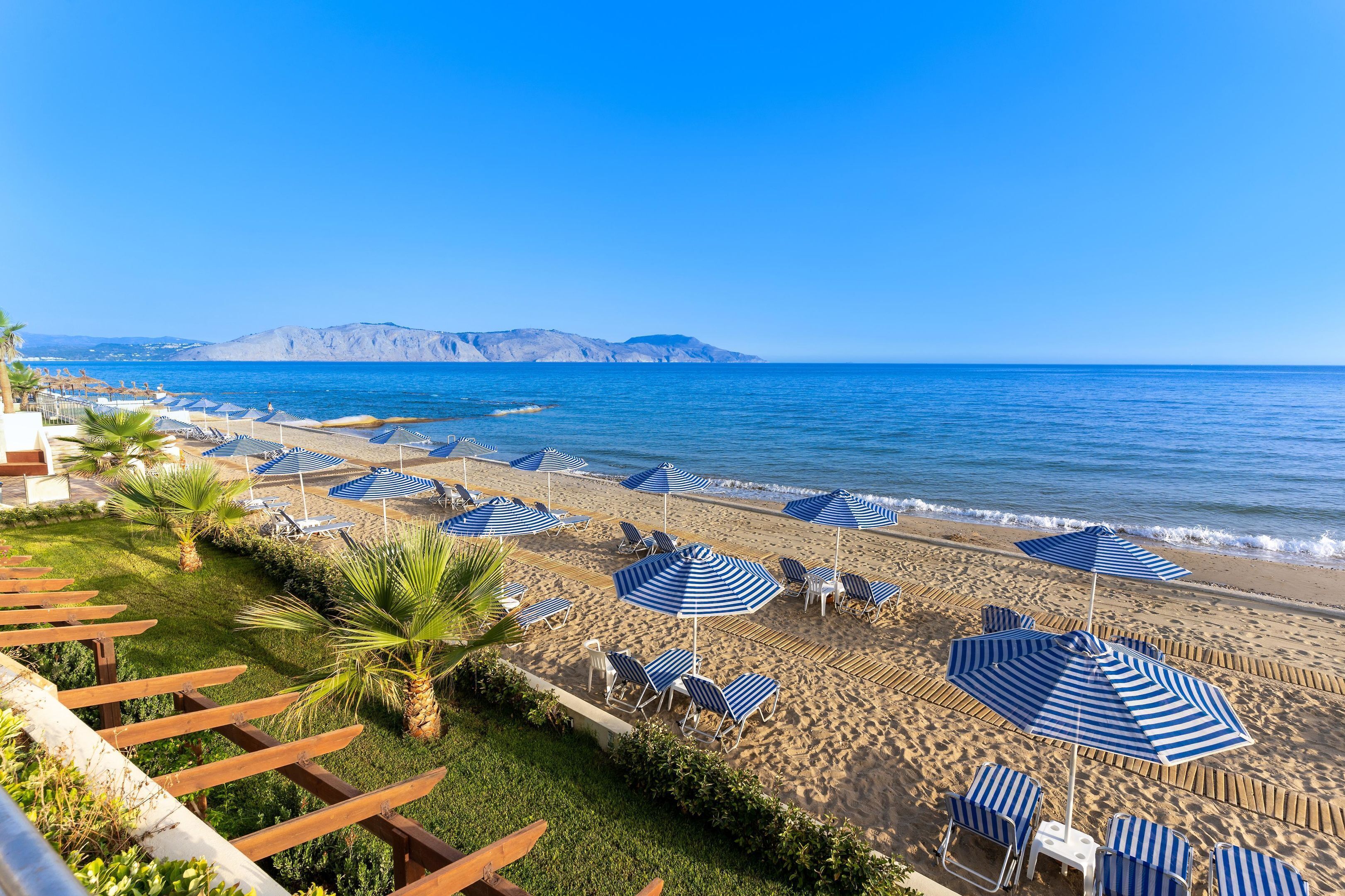 Una spiaggia con ombrelloni a righe blu e lettini, cielo sereno e una montagna sullo sfondo.