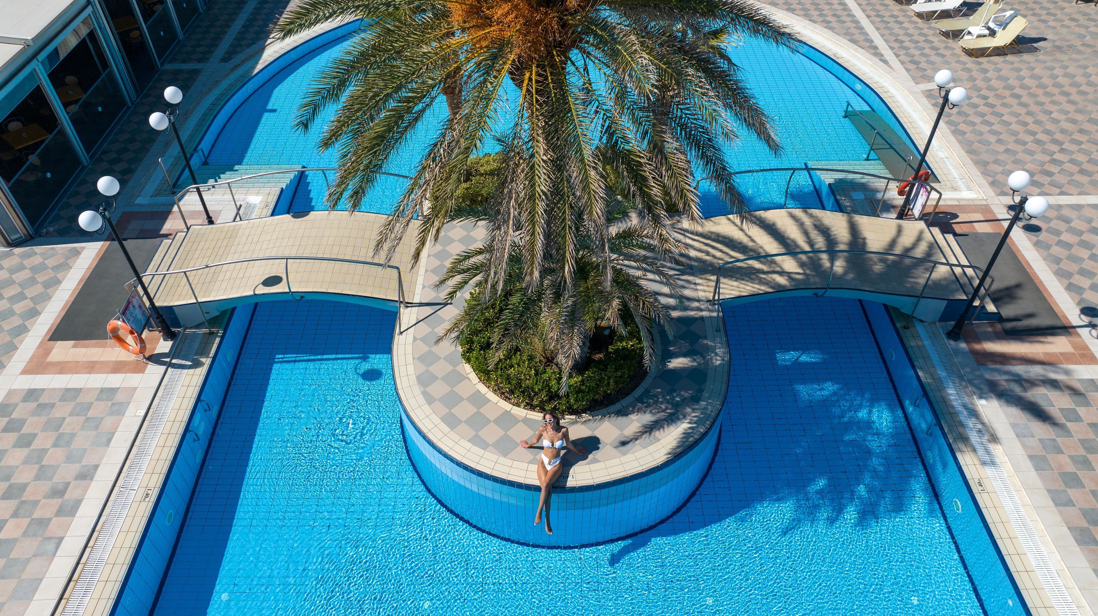 A circular pool with a palm tree in the center, surrounded by a tiled deck with lounge chairs and umbrellas.