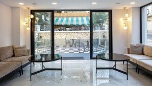 A modern interior with two sofas and two glass-top tables, a stone wall, and a view of an outdoor patio with a checkered floor and a striped awning. - Jo An Palace Hotel (Rethymno)