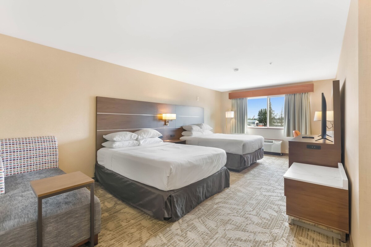 A hotel room with two beds, a desk, a chair, a TV, and a large window with curtains.