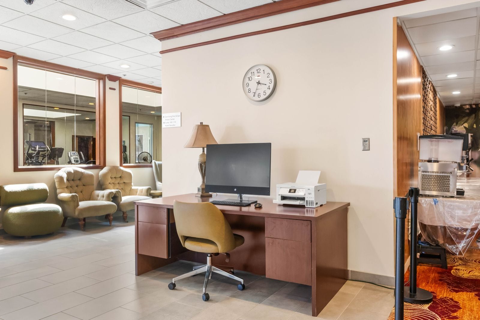A hotel room with a desk, computer, printer, and a clock on the wall.