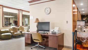 A hotel room with a desk, computer, printer, and a clock on the wall.