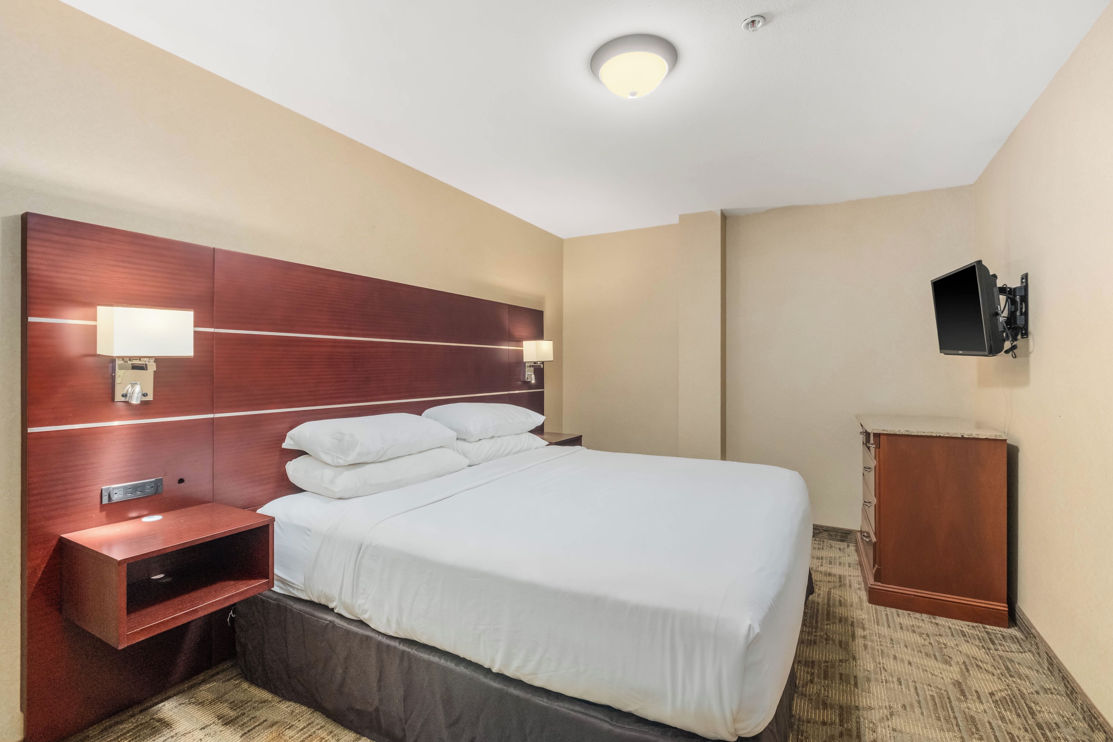 Une chambre d’hôtel avec un lit, des lampes de chevet, un téléviseur fixé au mur et une petite armoire en bois.