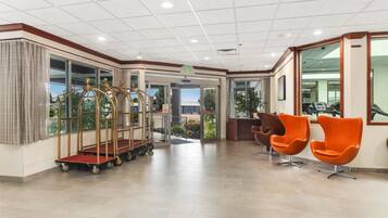 A hotel lobby with a reception desk, seating area with orange chairs, and a gold luggage cart.