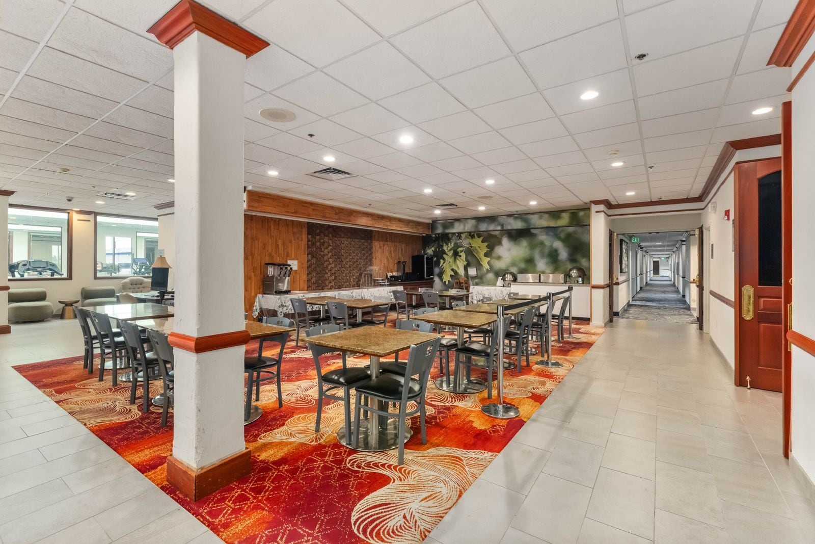 A hotel dining area with multiple tables and chairs, a kitchen area, and a hallway leading to an exit.