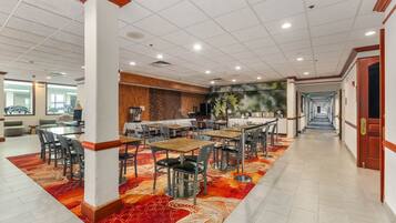 A hotel dining area with multiple tables and chairs, a kitchen area, and a hallway leading to an exit.