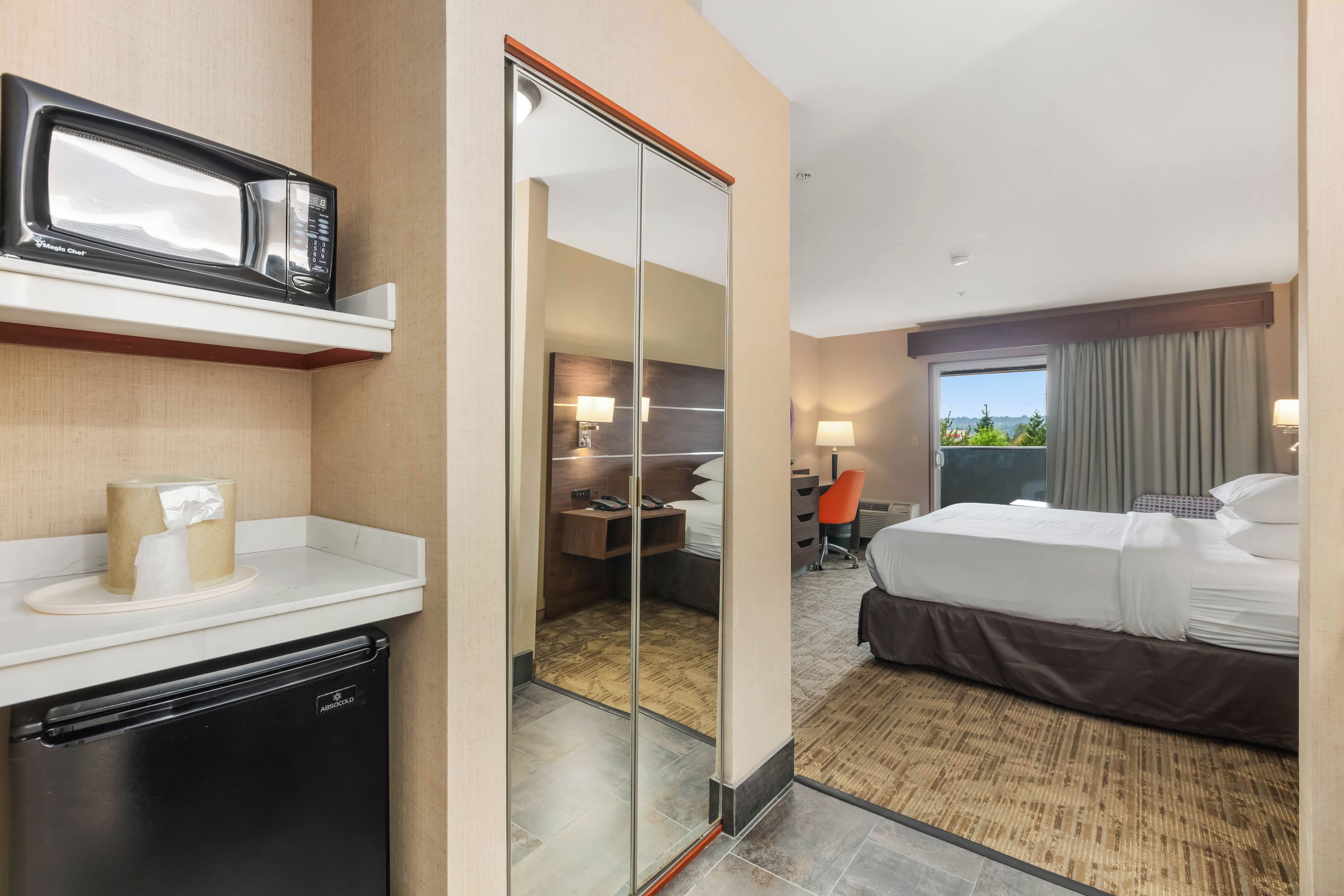 Una habitación de hotel con cama, escritorio, silla, microondas y espejo.