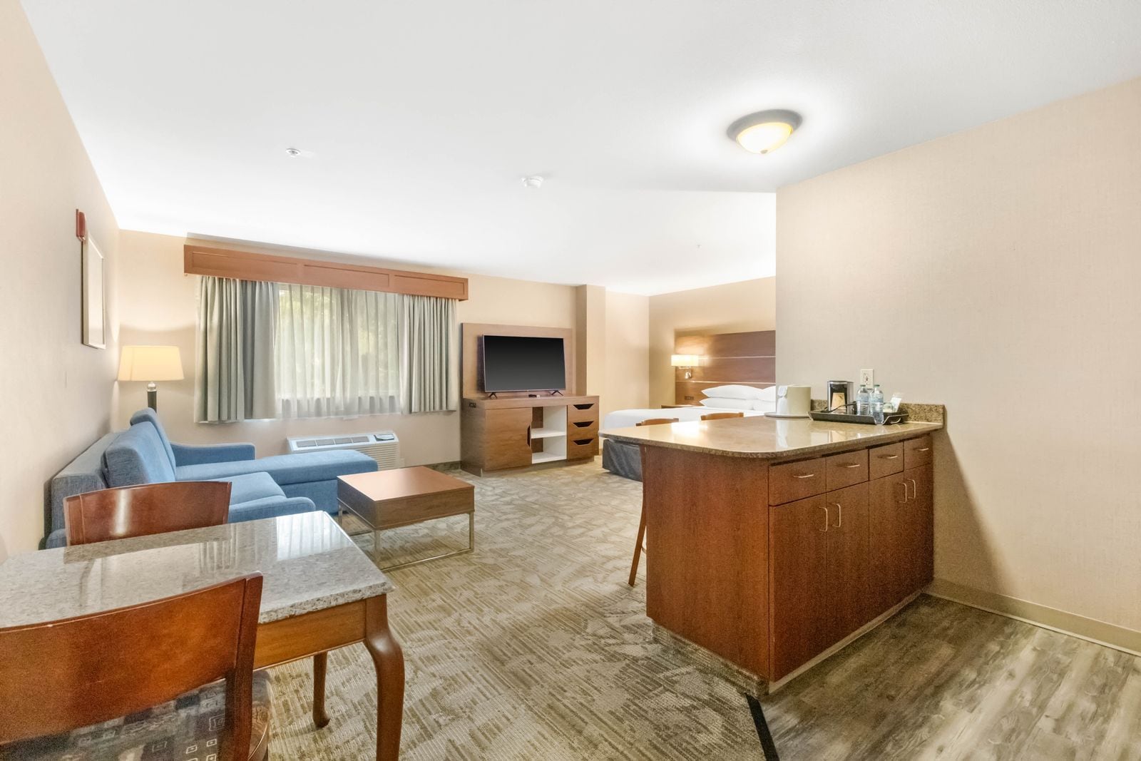 A hotel room with a living area, a TV, a sofa, a coffee table, a dining table, and a kitchen area with a counter and cabinets.