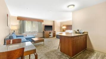 A hotel room with a living area, a TV, a sofa, a coffee table, a dining table, and a kitchen area with a counter and cabinets.