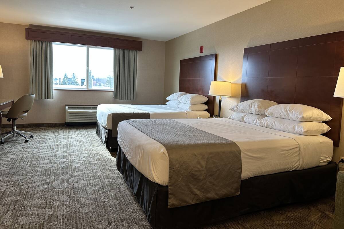 Habitación de hotel con dos camas, un escritorio, una silla, una ventana con cortinas y vistas a la ciudad.